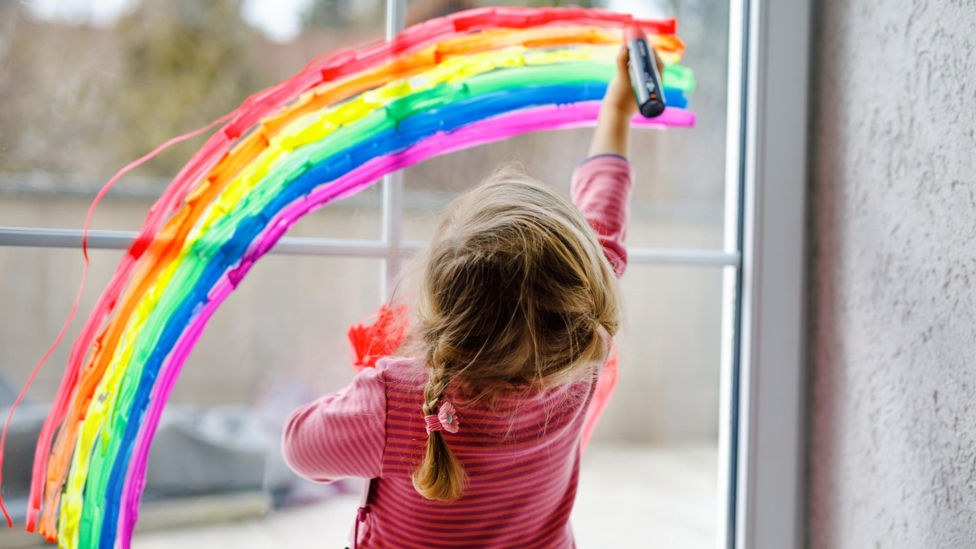 Ein Mädchen, welches an einer Fensterscheibe mit bunten Farben ein Regenbogen malt.