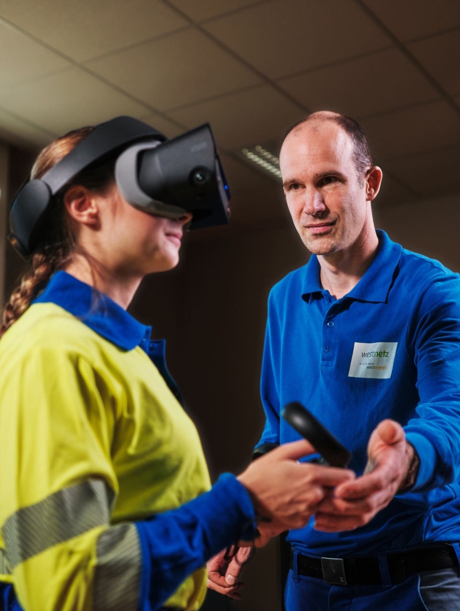 Ein Ausbilder leitet eine Auszubildende, die eine VR-Brille trägt, durch eine digitale Simulation eines Umspannwerks